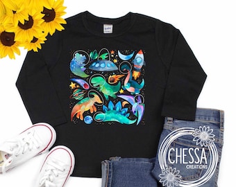 May include: A black long-sleeved shirt with a colorful graphic print of dinosaurs and spaceships. The shirt is laid flat on a white surface with a pair of white sneakers and blue jeans.
