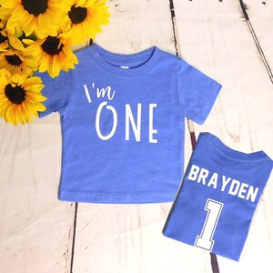 May include: Two blue t-shirts with white text. The first shirt says "I'm One" and the second shirt says "Brayden" with the number "1" underneath.