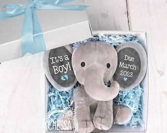 May include: A gray plush elephant toy in a white gift box with blue tissue paper. The elephant has two chalkboard ears with the text "It's a Boy!" and "Due March 2023".