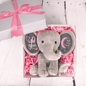K&ouml;nnte beinhalten: Ein graues Pl&uuml;sch-Elefantenspielzeug mit rosa Ohren und einem schwarzen Kreis auf jedem Ohr. Der Kreis auf dem linken Ohr sagt "Lilly Rose" in rosa Schrift. Der Kreis auf dem rechten Ohr sagt "Ich liebe dich bis zum Mond und zur&uuml;ck" in wei&szlig;er Schrift mit einem rosa Mond und Sternen. Der Elefant befindet sich in einer wei&szlig;en Geschenkbox mit rosa Seidenpapier.
