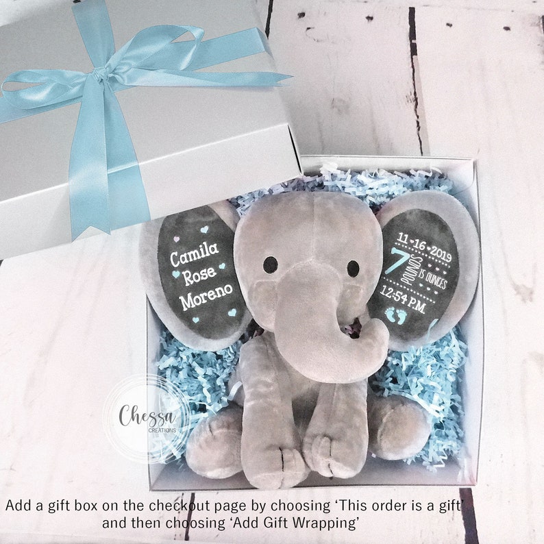 Puede incluir: Un juguete de peluche de elefante gris con detalles personalizados. El elefante est&aacute; sentado en una caja de regalo con papel de seda azul y blanco. Las orejas del elefante tienen el texto "Camila Rose Moreno" y la otra oreja tiene el texto "11-16-2019 7 pounds 12:54 PM".