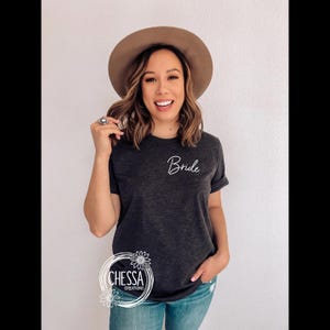 May include: Dark gray t-shirt with the word "Bride" in white script. The shirt has short sleeves and is paired with blue jeans. A brown hat is worn. The image also includes a logo with the text "CHESSA CREATIONS".