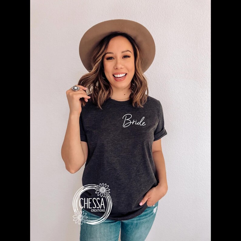 May include: Dark gray t-shirt with the word "Bride" in white script. The shirt has short sleeves and is paired with blue jeans. A brown hat is worn. The image also includes a logo with the text "CHESSA CREATIONS".