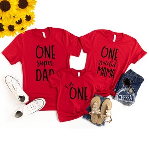 May include: Three red t-shirts with black text. The largest shirt says "One super Dad". The medium shirt says "One grateful Mama". The smallest shirt says "I'm One".