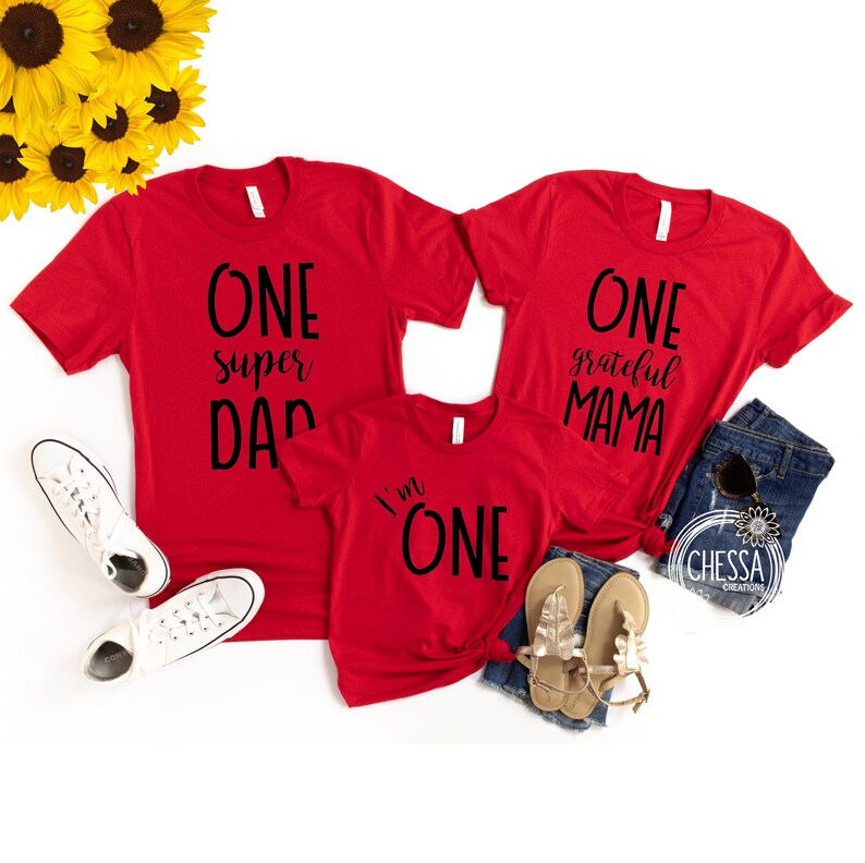 May include: Three red t-shirts with black text. The largest shirt says "One super Dad". The medium shirt says "One grateful Mama". The smallest shirt says "I'm One".