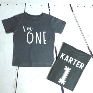 May include: Two gray t-shirts with white text. The first shirt says "I'm One" and the second shirt says "Karter 1".