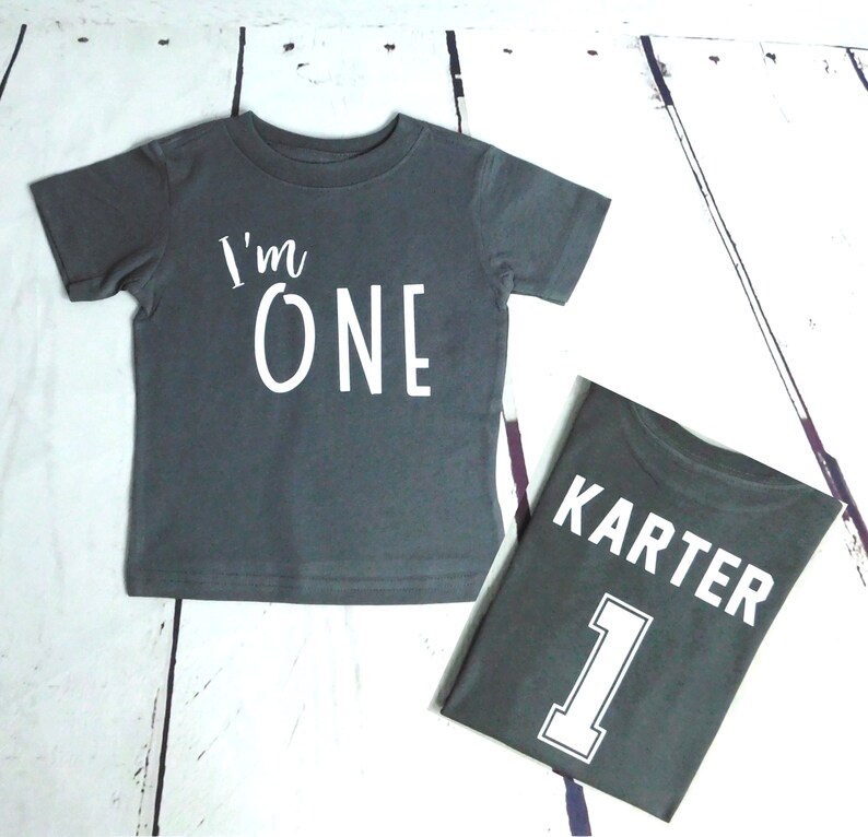 May include: Two gray t-shirts with white text. The first shirt says "I'm One" and the second shirt says "Karter 1".
