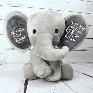 May include: Gray plush elephant stuffed animal with embroidered details. The ears are personalized with the name "Charlotte Ivy Aquino", birthdate "05-27-2019", weight "6 pounds 7 ounces", and time "4:17 AM".
