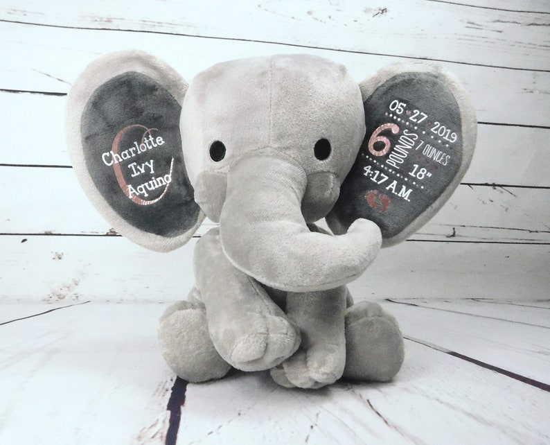 May include: Gray plush elephant stuffed animal with embroidered details. The ears are personalized with the name "Charlotte Ivy Aquino", birthdate "05-27-2019", weight "6 pounds 7 ounces", and time "4:17 AM".