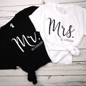 May include: Black and white t-shirts with the words "Mr." and "Mrs." printed in a cursive font. The date "Est. 04.28.2014" is printed below the names. The white shirt is tied in a knot at the bottom.