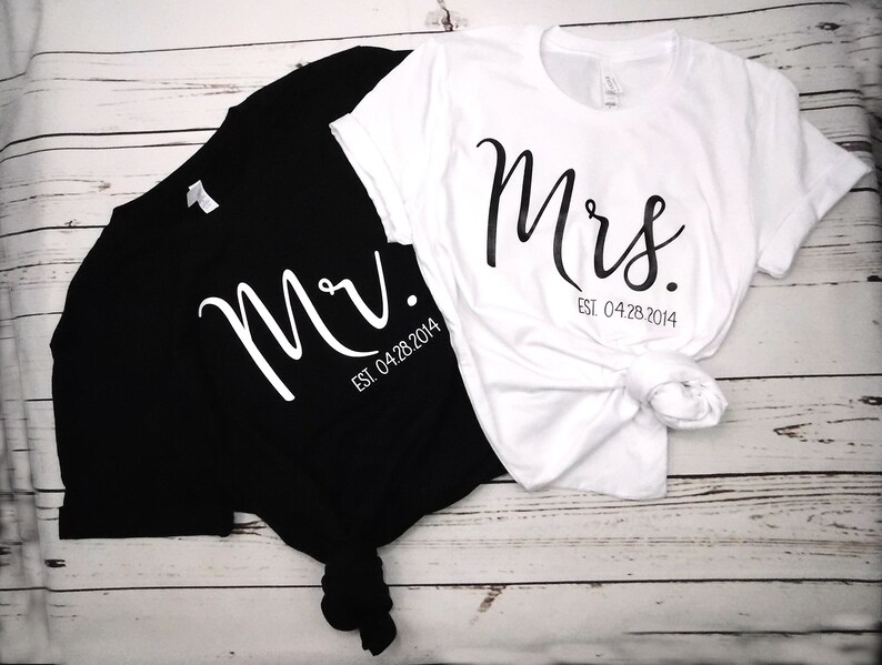 May include: Black and white t-shirts with the words "Mr." and "Mrs." printed in a cursive font. The date "Est. 04.28.2014" is printed below the names. The white shirt is tied in a knot at the bottom.