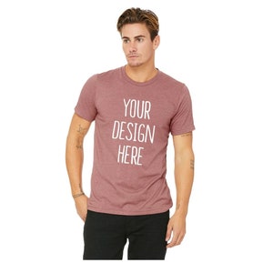 May include: A maroon crew neck t-shirt with the text "YOUR DESIGN HERE" in white. The short-sleeved shirt is made of a soft material and is suitable for custom designs. The model is wearing black pants.