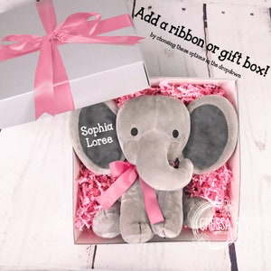 K&ouml;nnte beinhalten: Ein graues Pl&uuml;sch-Elefantenspielzeug mit rosa Schleife und dem Namen "Sophia Loree", gestickt auf dem Ohr, in einer wei&szlig;en Geschenkbox mit rosa Seidenpapier.