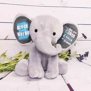 Puede incluir: Peluche de elefante gris de felpa con detalles bordados. Una oreja dice "Bruce Wayne" en texto azul y blanco. La otra oreja dice "9 lbs 1 oz, 17 de julio de 2018, 3:15 A.M., 21 pulgadas" en texto azul y blanco.