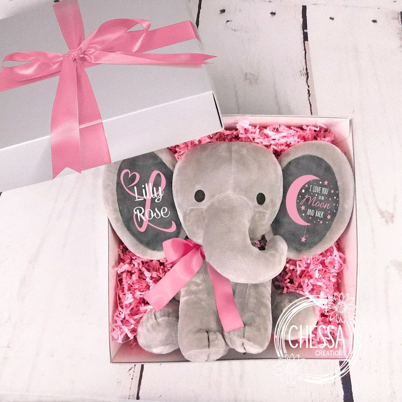 May include: A gray plush elephant stuffed animal with pink ribbon and ears. The ears have black felt circles with pink text. One ear says 'Lilly Rose' and the other ear says 'I love you to the Moon and Back'. The elephant is in a white gift box with pink tissue paper.