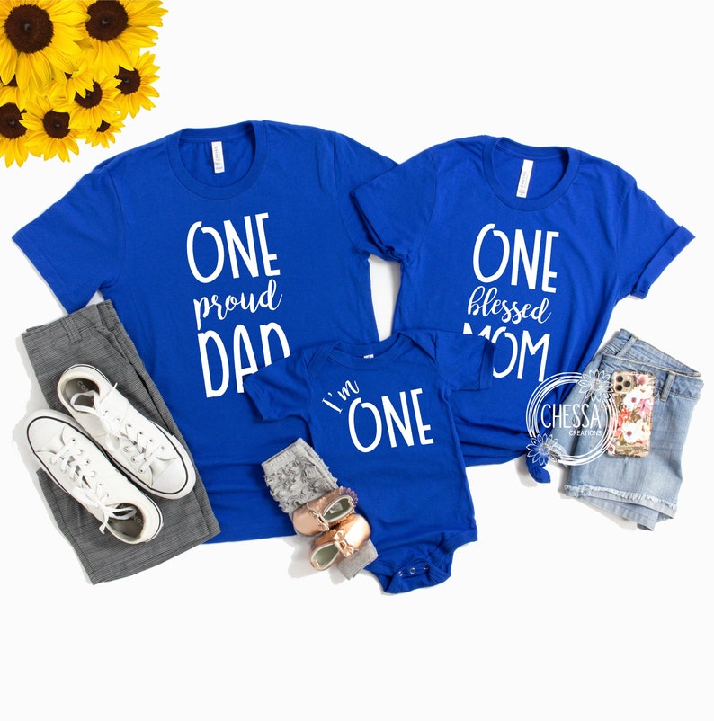 May include: A set of three blue t-shirts with white text. The largest shirt says "One proud Dad", the medium shirt says "One blessed Mom", and the smallest shirt says "I'm One".