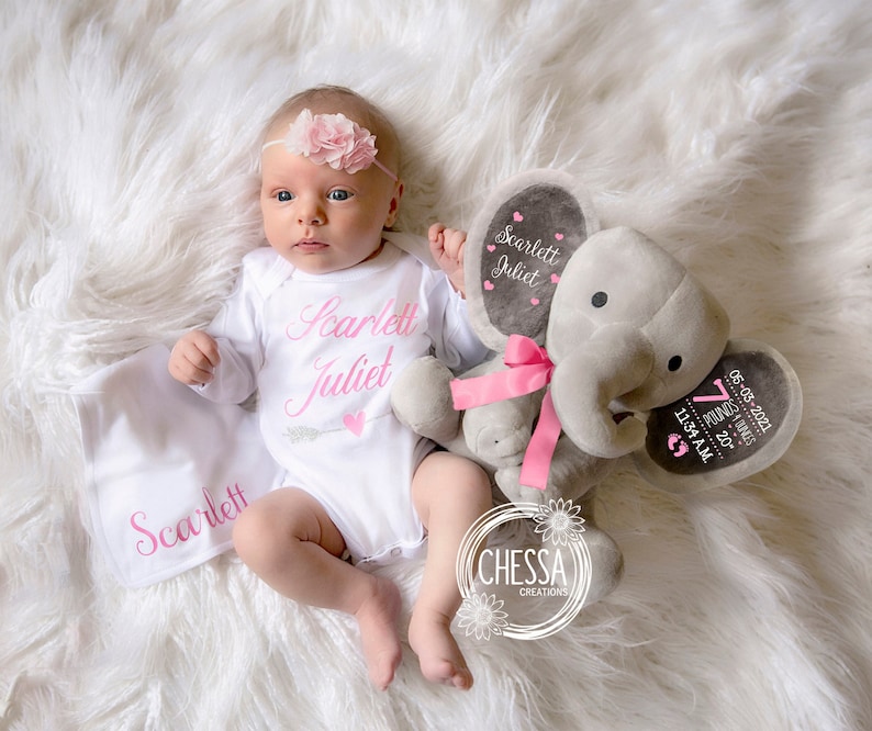 Puede incluir: Un peluche de elefante gris con un lazo rosa y una etiqueta personalizada con el nombre "Scarlett Juliet" y los detalles de nacimiento. El elefante est&aacute; al lado de un beb&eacute; que lleva un body blanco con el mismo nombre y una diadema de flores rosa.