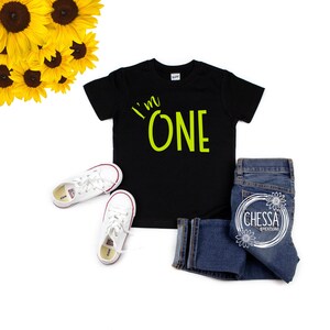 May include: A black t-shirt with the words "I'm One" in neon green lettering. The shirt is laid flat on a white background with a pair of white sneakers and blue jeans.