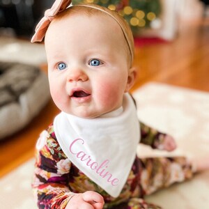 Puede incluir: Un beb&eacute; con un babero blanco con el nombre "Caroline" bordado en rosa. El babero se lleva sobre un mono de estampado floral. El beb&eacute; lleva una diadema rosa.
