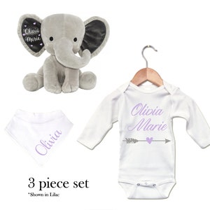 Puede incluir: Un conjunto de regalo para beb&eacute; de tres piezas que incluye un elefante de peluche gris, un body blanco de manga larga con el texto "Olivia Marie" y una flecha gris con un coraz&oacute;n, y un babero bandana blanco con el texto "Olivia" en morado.
