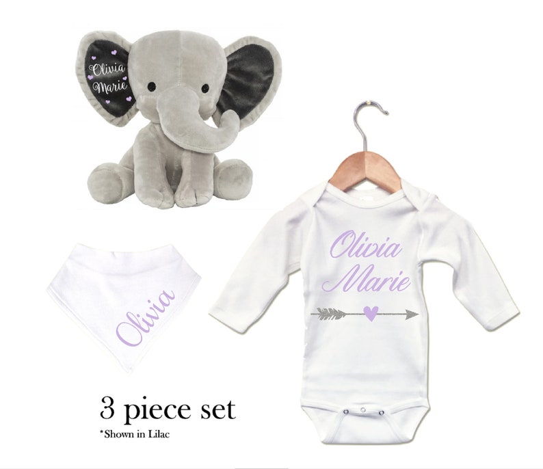 Puede incluir: Un conjunto de regalo para beb&eacute; de tres piezas que incluye un elefante de peluche gris, un body blanco de manga larga con el texto "Olivia Marie" y una flecha gris con un coraz&oacute;n, y un babero bandana blanco con el texto "Olivia" en morado.