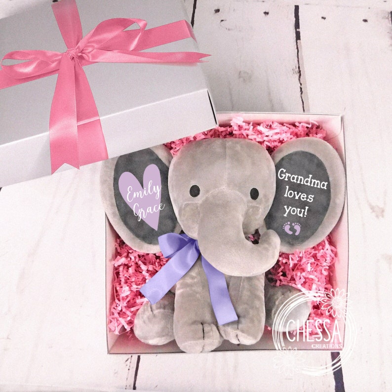 May include: A gray plush elephant toy with a purple ribbon around its neck. The elephant's ears are decorated with a purple heart that says "Emily Grace" and a black chalkboard that says "Grandma loves you!" The elephant is in a white gift box with pink tissue paper.