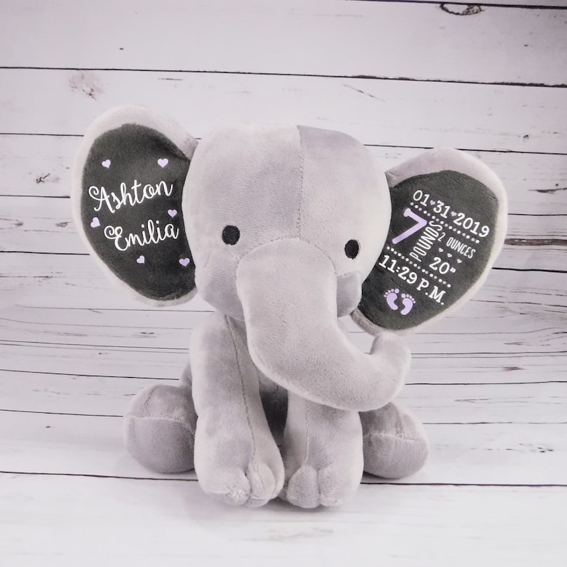 May include: Gray plush elephant stuffed animal with embroidered details. One ear says "Ashton Emilia" with pink hearts. The other ear says "01-31-2019 7 pounds 20 ounces 11:29 P.M." with pink footprints.