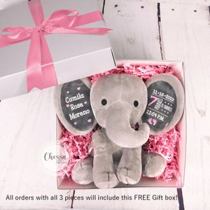 Puede incluir: Un elefante de peluche gris en una caja de regalo con papel de seda rosa. El elefante tiene dos c&iacute;rculos negros de fieltro para los ojos y un c&iacute;rculo rosa de fieltro para la nariz. El elefante sostiene una pizarra negra con el texto "Camila Rose Moreno" y otra pizarra con el texto "11-16-2019 7 libras 3 onzas 12:54 PM".