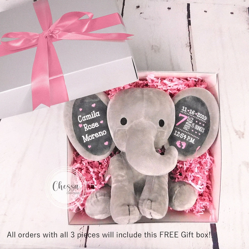 Puede incluir: Un elefante de peluche gris en una caja de regalo con papel de seda rosa. El elefante tiene dos c&iacute;rculos negros de fieltro para los ojos y un c&iacute;rculo rosa de fieltro para la nariz. El elefante sostiene una pizarra negra con el texto "Camila Rose Moreno" y otra pizarra con el texto "11-16-2019 7 libras 3 onzas 12:54 PM".