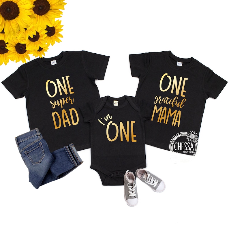 May include: A set of three black t-shirts with gold foil lettering. The shirts say "One super Dad", "One grateful Mama", and "I'm One".