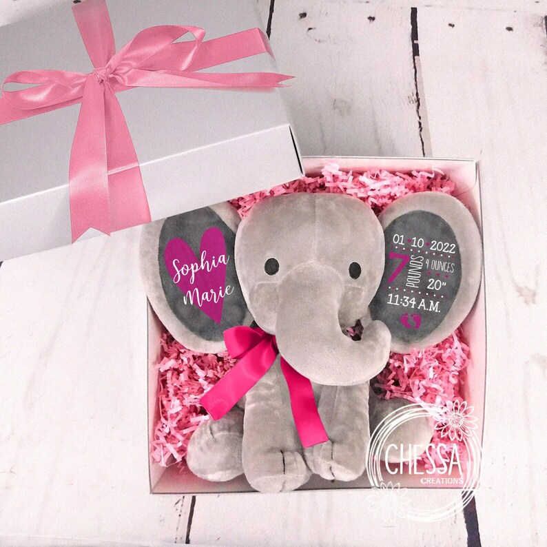 May include: A gray elephant plush toy with a pink bow and a heart-shaped tag that says "Sophia Marie". The elephant is in a white gift box with pink tissue paper. The box also includes a tag with the text "01.10.2022 7 pounds 9 ounces 20" 11:34 A.M."