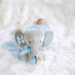 May include: A gray plush elephant with blue ribbon and embroidered details. The elephant's ears are personalized with the name "Noah James" and the date "03 03 2021" with the time "11:34 AM".