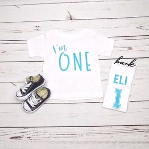May include: White onesie with blue text that says "I'm ONE" on the front and a white onesie with blue text that says "ELI 1" on the back.