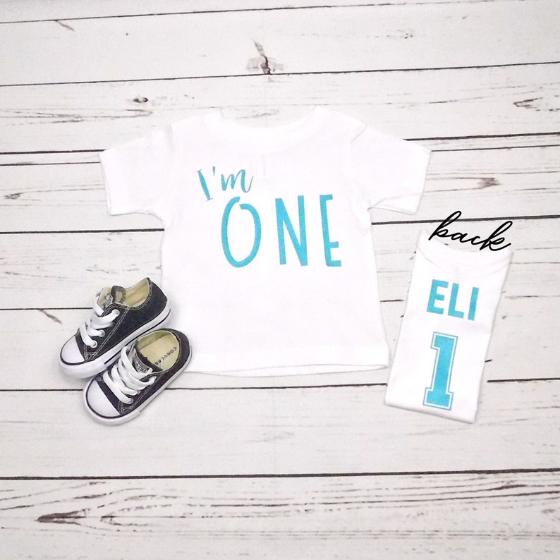 May include: White onesie with blue text that says "I'm ONE" on the front and a white onesie with blue text that says "ELI 1" on the back.