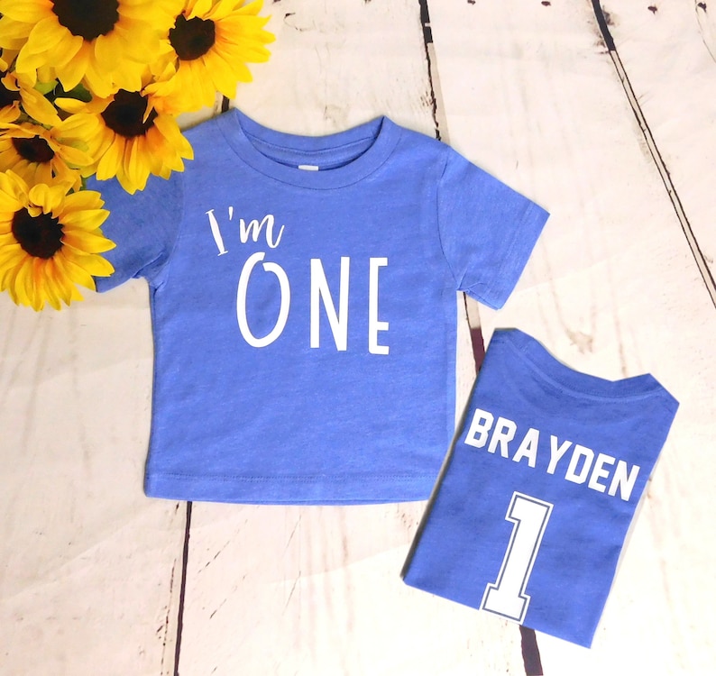 May include: Two blue t-shirts with white text. The first shirt says "I'm One" and the second shirt says "Brayden" with the number "1" underneath.