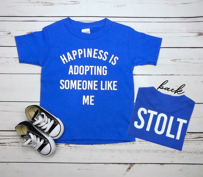 May include: A royal blue t-shirt with white text that reads "Happiness is adopting someone like me". The back of the shirt has the word "Stolt" in white text.