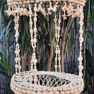 Może przedstawiać: Wiszący kwietnik wykonany z naturalnych muszli. Kwietnik ma kształt dzwonu z szeroką górą i wąskim dnem. Kwietnik jest ozdobiony frędzlami z muszli.