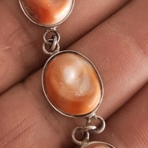 Puede incluir: Un colgante de plata con tres piedras ovaladas de color naranja y blanco. Las piedras están conectadas por eslabones de plata. El colgante se sostiene en una mano.