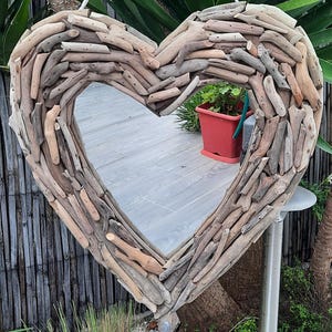 Può includere: Specchio a forma di cuore incorniciato con pezzi di legno trasportati dall'acqua. La cornice è realizzata con vari pezzi di legno stagionato nei toni del marrone e beige. Lo specchio riflette l'ambiente esterno.