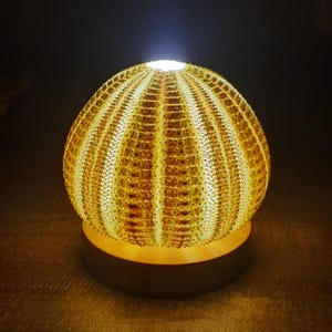 Op de afbeelding: Een decoratieve lamp met een uniek ontwerp. De bolvormige lampenkap is gemaakt van een getextureerd, lichtgekleurd materiaal, mogelijk een zee-egelschelp. Hij straalt een warme gloed uit en staat op een donkere houten voet. Geschikt voor nachtkastjes.