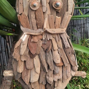 Peut inclure: Une décoration murale en forme de hibou, fabriquée à partir de nombreux morceaux de bois flotté brun clair et foncé. Le hibou présente deux yeux circulaires avec des centres métalliques et un corps texturé composé de bois disposé verticalement.