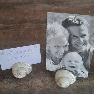 Könnte beinhalten: Ein Schwarzweißfoto einer Familie, einer Frau, eines Mannes und eines Babys, wird in einem Muschel-Bilderrahmen angezeigt. Das Foto ist 10x15 cm groß. Eine weiße Karte mit dem Text "Marriage d'Elena" wird in einem Muschel-Bilderrahmen angezeigt.