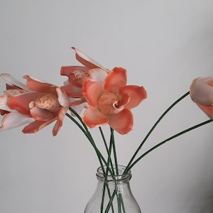 Op de afbeelding: Een heldere glazen vaas met kunstbloemen. De bloemen hebben perzik- en witte bloemblaadjes en groene stelen. De vaas is cilindrisch en staat tegen een witte achtergrond.