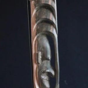 May include: A wooden sculpture of a stylized human figure with a carved face. The sculpture is dark brown and has a rough texture. It is standing on a black base.