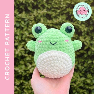 May include: A green crocheted frog amigurumi toy with white crocheted belly and black eyes. The frog is being held in a hand.