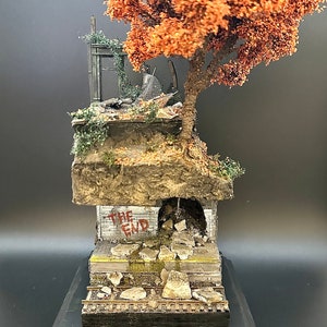 Puede incluir: Un diorama en miniatura que representa una ciudad en ruinas con un pequeño árbol creciendo fuera de las ruinas. El árbol tiene hojas marrones y las ruinas están hechas de hormigón gris. El diorama está colocado sobre una base negra. El texto "THE END" está pintado en una pared del diorama.