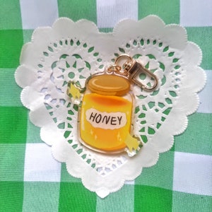 May include: A gold keychain charm shaped like a honey jar with a yellow lid and a clear body filled with honey. The jar has a white label with the word "HONEY" printed on it. The charm is attached to a gold split ring.