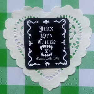 Puede incluir: Una pegatina cuadrada negra con texto blanco que dice "Jinx Hex Curse" sobre un gráfico de una sonrisa con dientes y un ojo en el centro. El texto "Magic with teeth" está debajo del gráfico. La pegatina está sobre un mantelito de encaje blanco.