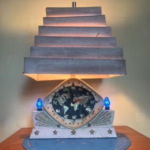 May include: A vintage table lamp with a world map clock face. The lamp has a stepped, rectangular shade and a base decorated with stars. Two blue light bulbs flank the clock. The clock face features gold-colored continents.