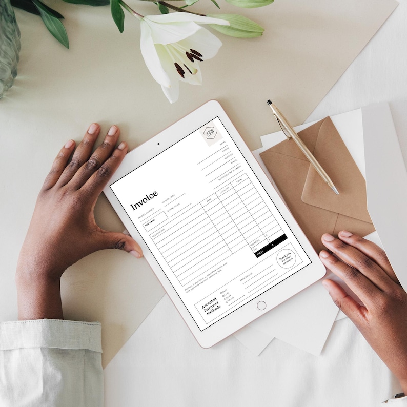May include: A white tablet displaying a black and white invoice template with the word "Invoice" at the top. A gold pen, a brown envelope, and white paper are next to the tablet. White flowers are in the background.